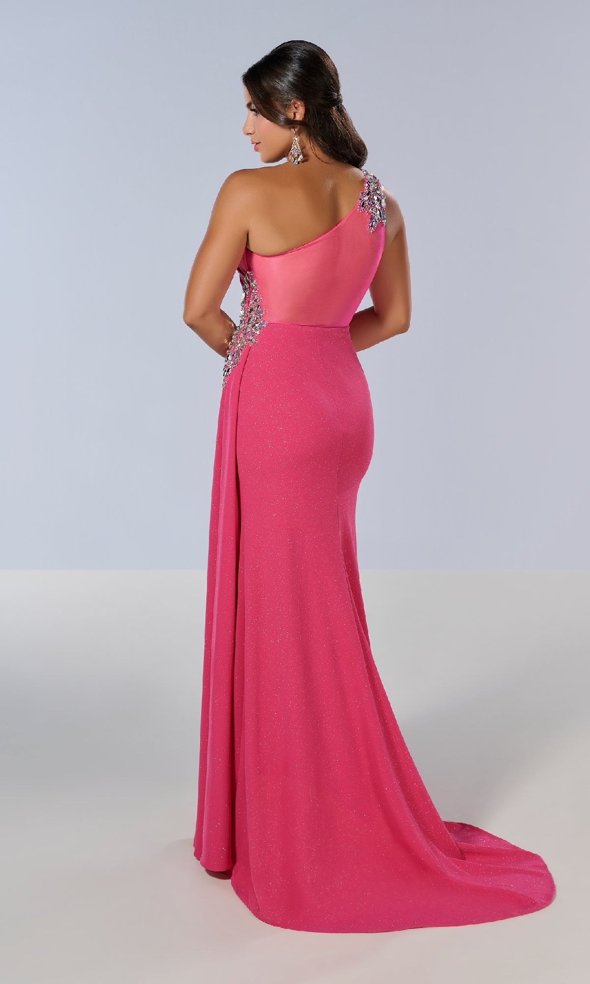 Woman in the Tiffany 16211 long prom dress, featuring a floor-length, one-shoulder design with jeweled embellishments, stands with her back to the camera against a plain background.